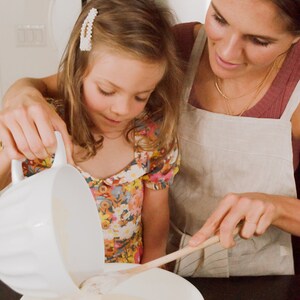 May include: A young girl pours batter into a bowl while a woman stirs the batter with a wooden spoon. The girl is wearing a floral dress and the woman is wearing a linen apron.