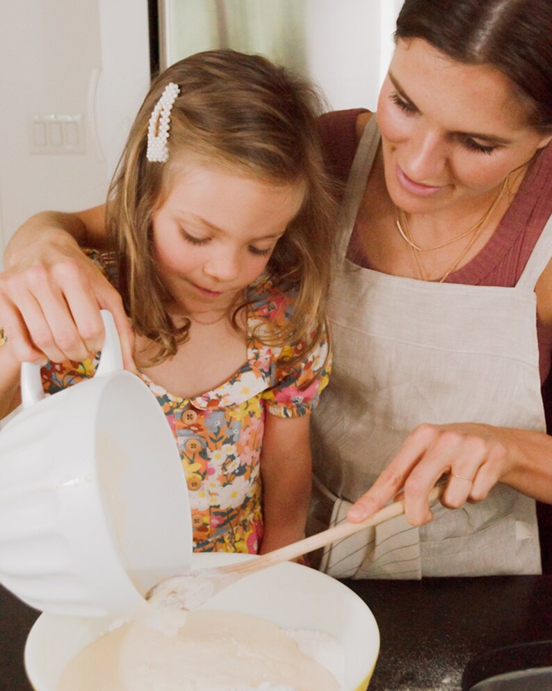 May include: A young girl pours batter into a bowl while a woman stirs the batter with a wooden spoon. The girl is wearing a floral dress and the woman is wearing a linen apron.