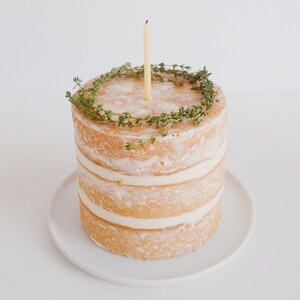 May include: A simple, three-tiered cake with a single candle on top. The cake is decorated with a ring of fresh thyme.