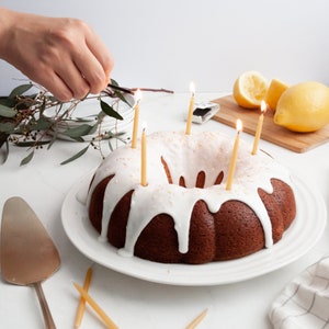 May include: A white frosted bundt cake with yellow candles on a white plate. The cake is decorated with white sprinkles. A hand is lighting the candles with a match.