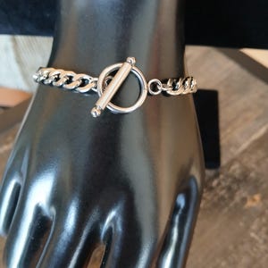 May include: A silver-colored curb chain bracelet with a toggle clasp. The bracelet is displayed on a black mannequin hand. The clasp features a circular ring and a cylindrical bar. The chain links are thick and interlocked.