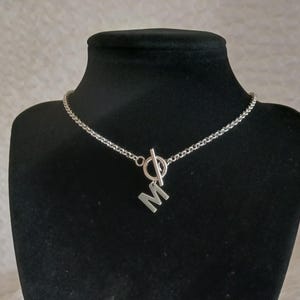 May include: Silver-toned necklace with a toggle clasp and a pendant featuring the letter "M". The chain has a textured, woven design. The necklace is displayed on a black mannequin bust against a neutral background.