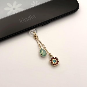 Op de afbeelding: Een goudkleurige bedel met een druppelvormige groene edelsteen en een bloem met rode en blauwe accenten, bevestigd aan een zwarte Kindle e-reader. De e-reader heeft het woord "kindle" erop gedrukt. De bedel is een decoratief accessoire.