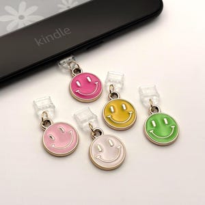 May include: A collection of colorful smiley face charms in pink, yellow, green, and light pink. Each charm has a gold-colored border and a clear plastic attachment. The word "kindle" is visible on a black electronic device.