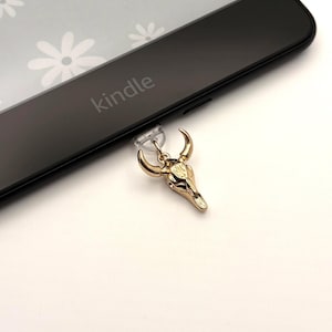 May include: A gold-colored bull skull charm with a clear suction cup, resting on a black Kindle device. The Kindle displays a floral pattern on its screen. The word "kindle" is printed on the device.