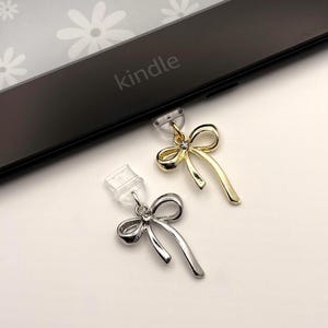 May include: Two bow-shaped charms, one gold and one silver, are displayed. The gold charm has a small gem at the center. The charms are attached to clear plastic pieces. The word "kindle" is visible on a black electronic device.