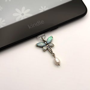 May include: A dragonfly-shaped charm with opalescent wings and a pearl drop hangs from a Kindle e-reader. The dragonfly is detailed with small crystals and a bronze-colored metal frame. The e-reader is black with the word "kindle" printed on it.