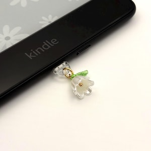 May include: A clear, flower-shaped charm with a gold-coloured loop and green leaf accents, attached to a black Kindle device. The word "kindle" is printed on the device. The charm has a small, gold-coloured centre.