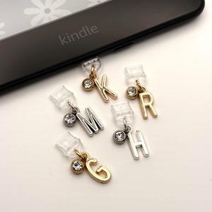 Puede incluir: Un conjunto de dijes decorativos para Kindle con las letras K, R, M, G y H. Cada dije tiene un pequeño clip transparente y un acento de pedrería brillante. Las letras son en tonos dorados y plateados, con un Kindle en el fondo.