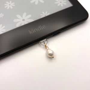 May include: A black Kindle e-reader with the word "kindle" printed on the front. A small, clear charm with a gold-coloured chain and a white pearl hangs from the bottom of the device.