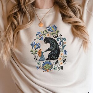 May include: Beige sweatshirt featuring a black bear and a child surrounded by a floral design in blue, green, and yellow. The graphic is centered on the front of the long-sleeved top. The sweatshirt is made of a soft material.