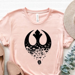 May include: A pink t-shirt with a black Rebel Alliance symbol made up of smaller symbols. The symbol is centered on the shirt. The shirt is laid flat on a white surface with two pairs of shoes and a hat.