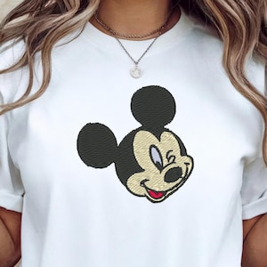 Puede incluir: Camiseta blanca con un bordado de la cara de Mickey Mouse. El dise&ntilde;o incluye orejas negras, una cara beige y una boca roja. La camiseta tiene mangas cortas y cuello redondo.