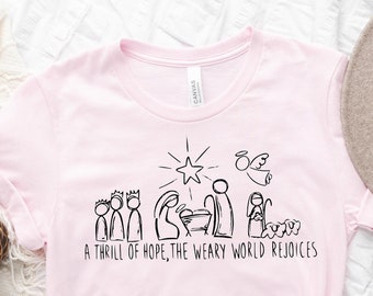 Op de afbeelding: Een lichtroze t-shirt met een zwarte lijntekening van een kerststal, een ster, een engel en de tekst "A thrill of hope, the weary world rejoices". Het t-shirt ligt op een wit, getextureerd oppervlak.