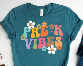 Könnte beinhalten: Türkisfarbenes T-Shirt mit dem Schriftzug "Pre-K Vibes" in einem Retro-Schriftstil, verziert mit bunten Blumen. Das Shirt wird mit blauen Jeans und weißen Turnschuhen kombiniert. Ein Holztablett mit einer Pflanze, einer Kamera, einer Uhr, einer Brille und einer Geldbörse ist ebenfalls im Bild.