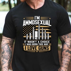 May include: A black short-sleeved t-shirt featuring a graphic print with the text I'M AMMOSEXUAL, IT WASN'T A CHOICE I WAS BORN THIS WAY, and I LOVE GUNS. The design includes a stylized American flag background and an illustration of various bullets.
