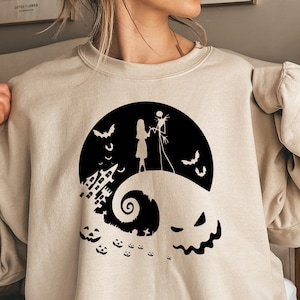 K&ouml;nnte beinhalten: Ein beiger Pullover mit einem schwarzen Grafikdruck von Jack Skellington und Sally aus Nightmare Before Christmas. Die Grafik zeigt sie auf einem H&uuml;gel stehend, im Hintergrund eine gruselige Halloween-Stadt.