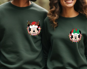 Puede incluir: Dos sudaderas verde oscuro con diseños navideños de Mickey y Minnie Mouse. Una sudadera presenta a Mickey con cuernos de reno y un gorro de Papá Noel, rodeado de luces navideñas. La otra tiene a Minnie con un lazo verde y un gorro de Papá Noel, también con luces.