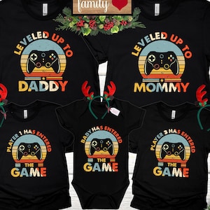 May include: A set of four black t-shirts with a retro video game theme. The shirts feature a vintage-style joystick graphic with the text "Leveled Up To" and "Daddy", "Mommy", "Player 1 Has Entered The Game", and "Player 3 Has Entered The Game".