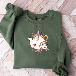 May include: A dark green sweatshirt featuring a cartoon illustration of a teapot and teacup. The design is in shades of gold, white, and purple, with star accents. The sweatshirt has long sleeves and a crew neck.