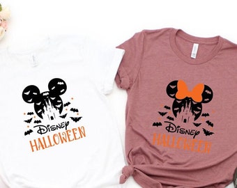 May include: Two t-shirts with a Halloween theme. One is white, the other is mauve. Both feature a black Mickey Mouse head with a castle, bats, and the words "Disney Halloween" in orange. The mauve shirt has an orange bow.