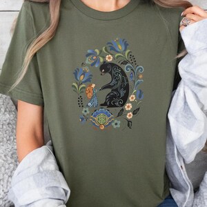 May include: Olive green t-shirt featuring a black bear and a figure with floral and foliate design. The bear and figure are surrounded by a wreath of blue, orange, and white flowers. The shirt is short-sleeved.