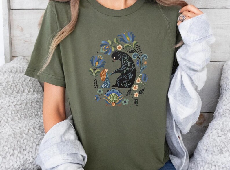 May include: Olive green t-shirt featuring a black bear and a figure with floral and foliate design. The bear and figure are surrounded by a wreath of blue, orange, and white flowers. The shirt is short-sleeved.