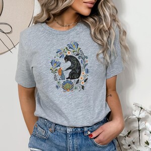 May include: A light gray t-shirt featuring a colorful floral design with a black bear and a smaller figure. The design is centered on the shirt and surrounded by blue, green, and orange floral elements. The shirt is made of a soft material.
