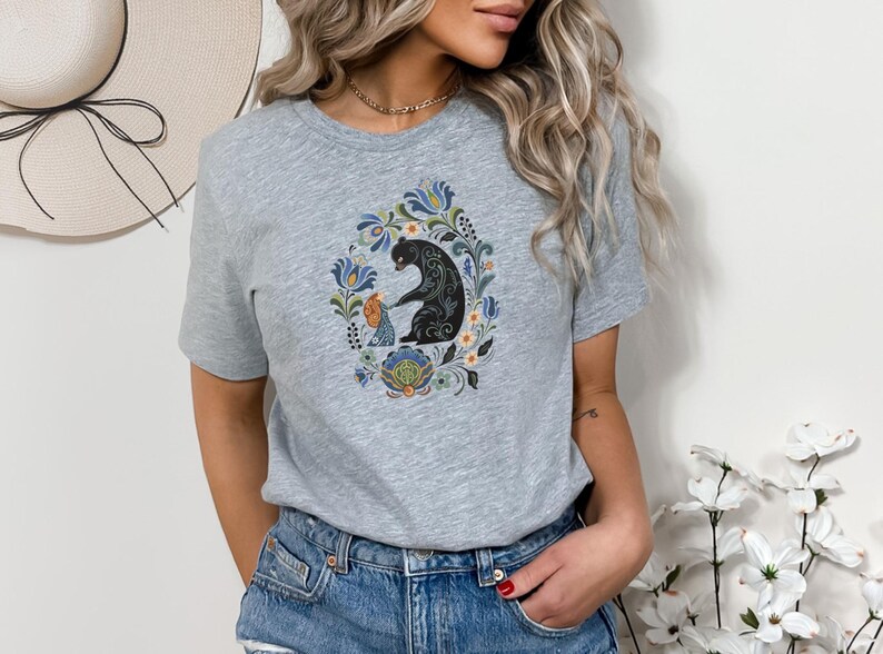 May include: A light gray t-shirt featuring a colorful floral design with a black bear and a smaller figure. The design is centered on the shirt and surrounded by blue, green, and orange floral elements. The shirt is made of a soft material.