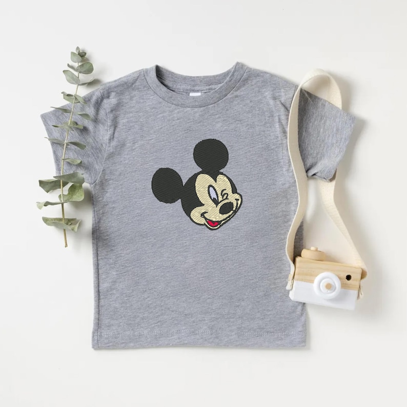 Puede incluir: Una camiseta gris con un bordado de la cara de Mickey Mouse. El dise&ntilde;o incluye orejas negras, una cara beige y una boca roja. La camiseta de manga corta parece estar hecha de un material suave. A la derecha hay una peque&ntilde;a c&aacute;mara de juguete con una correa blanca.
