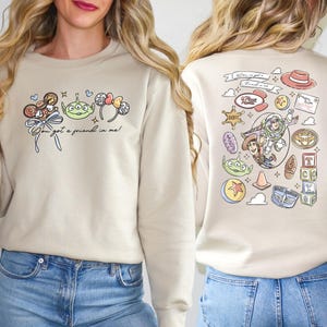 Pode incluir: Camisola bege com gr&aacute;ficos de desenhos animados coloridos. A frente apresenta um design de doces, alien&iacute;genas e orelhas de rato com o texto "You've got a friend in me!". A parte de tr&aacute;s tem v&aacute;rias ilustra&ccedil;&otilde;es de personagens.