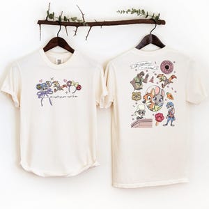 Pode incluir: Duas camisetas creme s&atilde;o exibidas. A frente de uma camiseta apresenta um design de desenho animado com uma c&acirc;mera, donuts e o texto "you can do anything you want to do". A parte de tr&aacute;s da outra camiseta tem um design de desenho animado colorido com v&aacute;rios personagens.