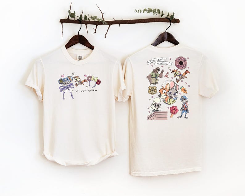 Pode incluir: Duas camisetas creme s&atilde;o exibidas. A frente de uma camiseta apresenta um design de desenho animado com uma c&acirc;mera, donuts e o texto "you can do anything you want to do". A parte de tr&aacute;s da outra camiseta tem um design de desenho animado colorido com v&aacute;rios personagens.
