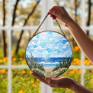 May include: Round stained glass art piece depicting a beach scene with blue sky, white clouds, and ocean waves. The artwork features a sandy path, green grass, and is framed with a black metal chain for hanging.