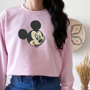 Puede incluir: Sudadera rosa claro con un bordado de la cara de Mickey Mouse. El ic&oacute;nico personaje de dibujos animados tiene orejas negras, cara amarilla y boca roja. La sudadera tiene mangas largas y cuello redondo.
