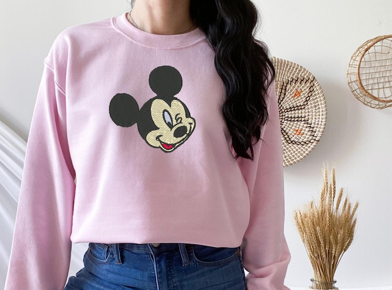 Puede incluir: Sudadera rosa claro con un bordado de la cara de Mickey Mouse. El ic&oacute;nico personaje de dibujos animados tiene orejas negras, cara amarilla y boca roja. La sudadera tiene mangas largas y cuello redondo.