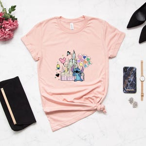 May include: A pink t-shirt with a graphic of Stitch holding balloons in front of a pink castle. The castle has a Mickey Mouse head on top and there are other Disney characters and icons around it.