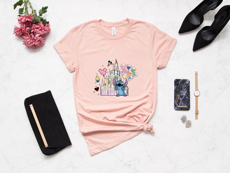 May include: A pink t-shirt with a graphic of Stitch holding balloons in front of a pink castle. The castle has a Mickey Mouse head on top and there are other Disney characters and icons around it.
