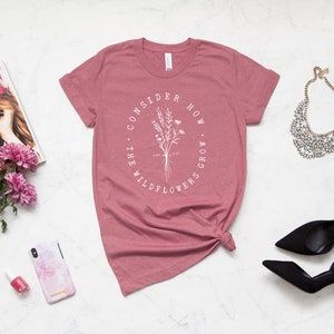 May include: A dusty rose t-shirt with the text "CONSIDER HOW THE WILDFLOWERS GROW" in a white oval around a floral illustration. The shirt is styled with accessories including a magazine, flowers, and heels.