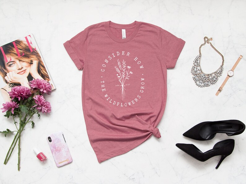 May include: A dusty rose t-shirt with the text "CONSIDER HOW THE WILDFLOWERS GROW" in a white oval around a floral illustration. The shirt is styled with accessories including a magazine, flowers, and heels.