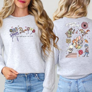 Pode incluir: Camisola cinza claro com ilustra&ccedil;&otilde;es de desenhos animados coloridos na frente e atr&aacute;s. A frente apresenta um personagem de rato, um guaxinim e detalhes florais com o texto "You can be anything you wish to be". A parte de tr&aacute;s tem v&aacute;rios personagens de desenhos animados.