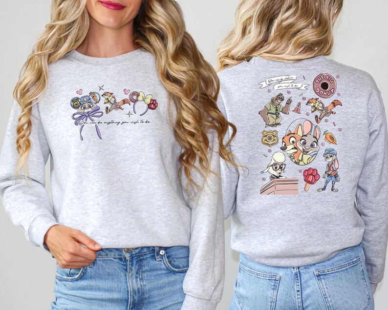 Pode incluir: Camisola cinza claro com ilustra&ccedil;&otilde;es de desenhos animados coloridos na frente e atr&aacute;s. A frente apresenta um personagem de rato, um guaxinim e detalhes florais com o texto "You can be anything you wish to be". A parte de tr&aacute;s tem v&aacute;rios personagens de desenhos animados.