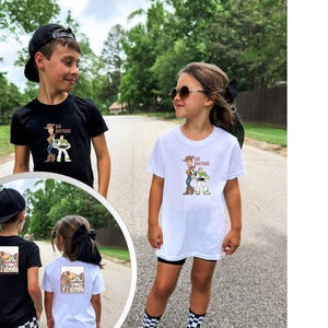 May include: Two children wearing black and white t-shirts featuring a cartoon graphic of Woody and Buzz Lightyear from Toy Story. The shirts have the text "Big Brother" on the front. The children are standing on a paved road.