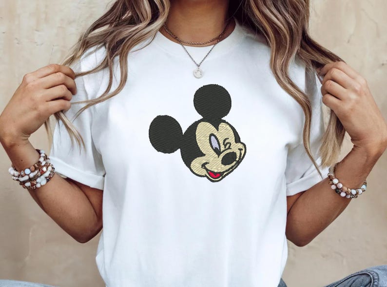 Puede incluir: Camiseta blanca con un bordado de la cara de Mickey Mouse. El dise&ntilde;o incluye orejas negras, una cara beige y una boca roja. La camiseta tiene mangas cortas y cuello redondo.