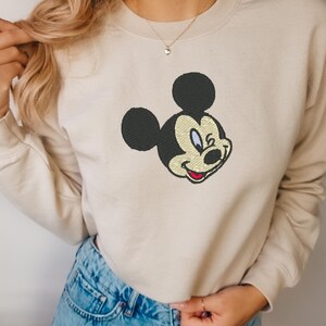 Puede incluir: Sudadera beige con un bordado de la cara de Mickey Mouse. El dise&ntilde;o incluye orejas negras, una cara color crema y una boca roja. La sudadera tiene mangas largas y cuello redondo.