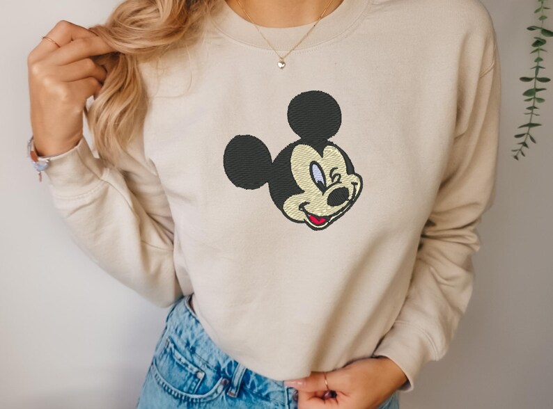 Puede incluir: Sudadera beige con un bordado de la cara de Mickey Mouse. El dise&ntilde;o incluye orejas negras, una cara color crema y una boca roja. La sudadera tiene mangas largas y cuello redondo.
