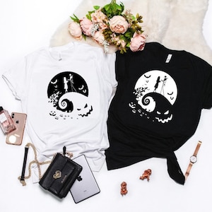 K&ouml;nnte beinhalten: Zwei T-Shirts mit einem Schwarz-Wei&szlig;-Motiv von Jack Skellington und Sally aus dem Film Nightmare Before Christmas. Das Motiv zeigt sie auf einem H&uuml;gel stehend mit einem Mond und Flederm&auml;usen im Hintergrund. Ein Shirt ist wei&szlig; und das andere ist schwarz.