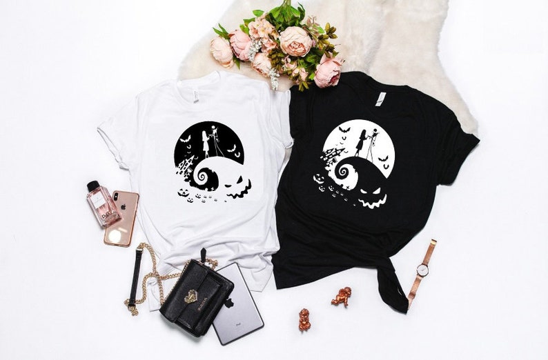 K&ouml;nnte beinhalten: Zwei T-Shirts mit einem Schwarz-Wei&szlig;-Motiv von Jack Skellington und Sally aus dem Film Nightmare Before Christmas. Das Motiv zeigt sie auf einem H&uuml;gel stehend mit einem Mond und Flederm&auml;usen im Hintergrund. Ein Shirt ist wei&szlig; und das andere ist schwarz.