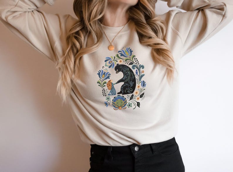 May include: Beige sweatshirt featuring a black bear and a child surrounded by a floral design in blue, green, and yellow. The graphic is centered on the front of the long-sleeved top. The sweatshirt is made of a soft material.