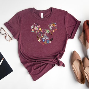 May include: A maroon t-shirt with a Mickey Mouse head design made up of various Disney characters and icons. The shirt is laid flat on a white surface.